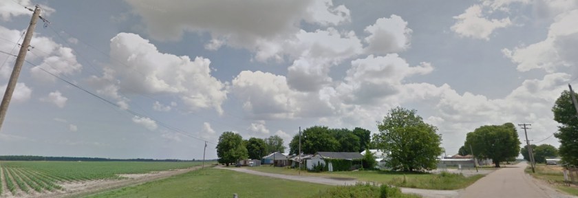Paved and gravel roads. Farm fields and small houses. Blue sky with clouds on a sunny day.