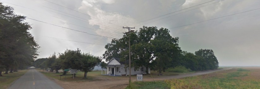 Paved and gravel roads. Farm fields and a small church. Cloudy skies.