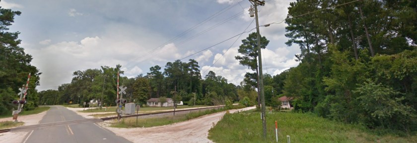 Paved and gravel roads. Train crossing and small homes among trees. Blue skies.
