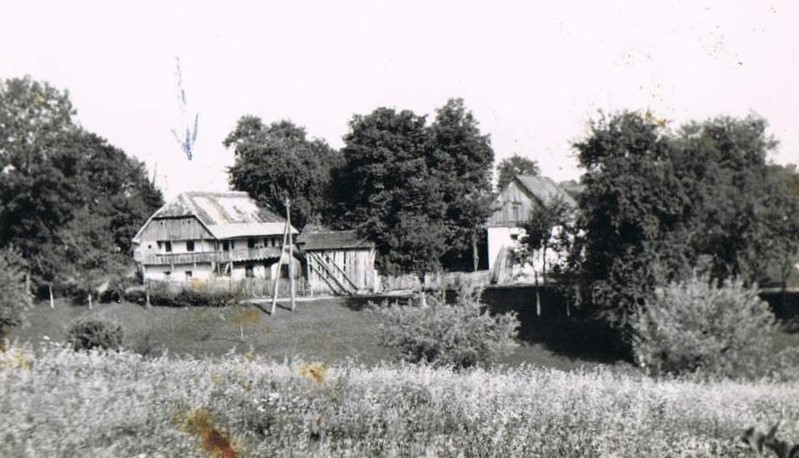 A historical black and white photograph of a rustic farmhouse surrounded by trees and tall grass, likely from an earlier era.