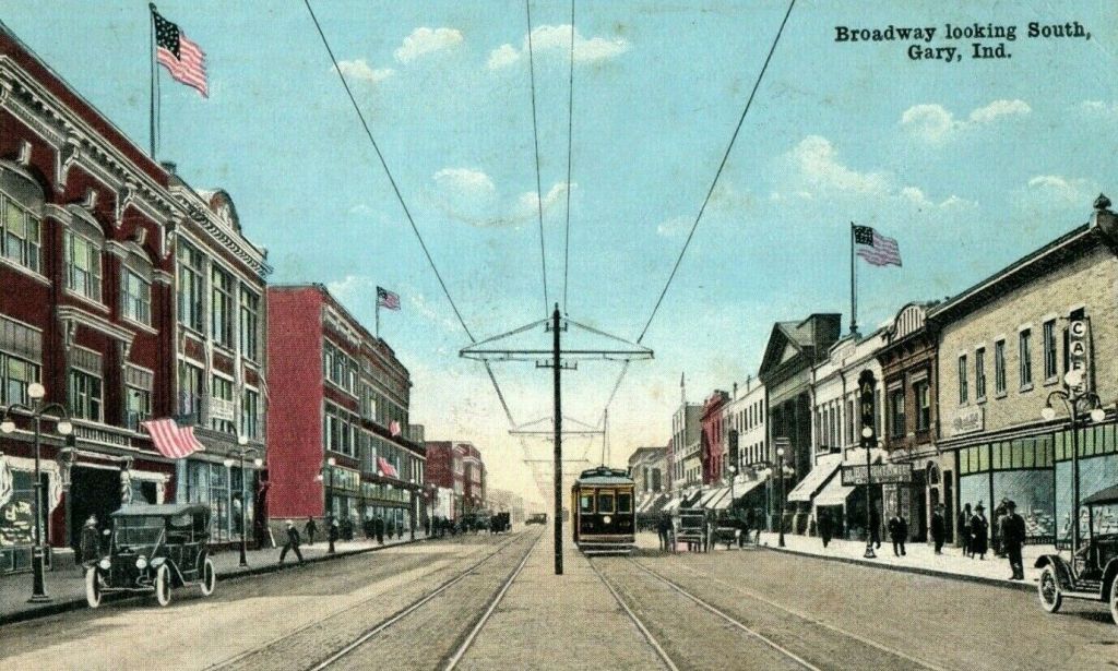 A vintage postcard depicting Broadway looking south in Gary, Indiana, with American flags adorning the buildings, early 20th-century vehicles on the street, and a streetcar on the tracks.