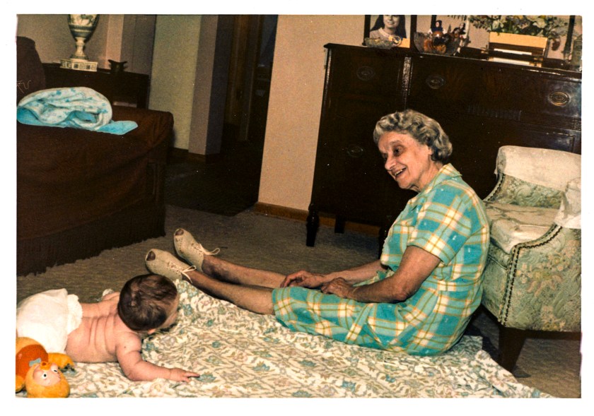 An elderly woman in a green and yellow checkered dress sits on a patterned rug, smiling at a baby lying on their stomach nearby. The room contains a wooden dresser and a decorative chair.