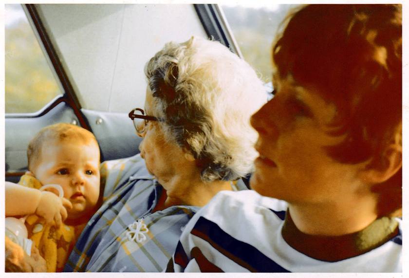 A baby being held by an elderly woman, with a young woman seated nearby, all inside a car. The scene captures a moment of family connection and travel.